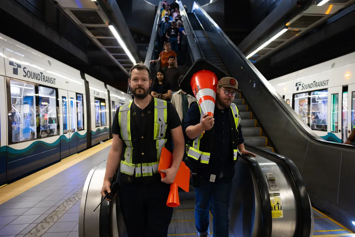 Two workers wearing safety vests step off an escalator in a light rail station carrying construction cones and signage