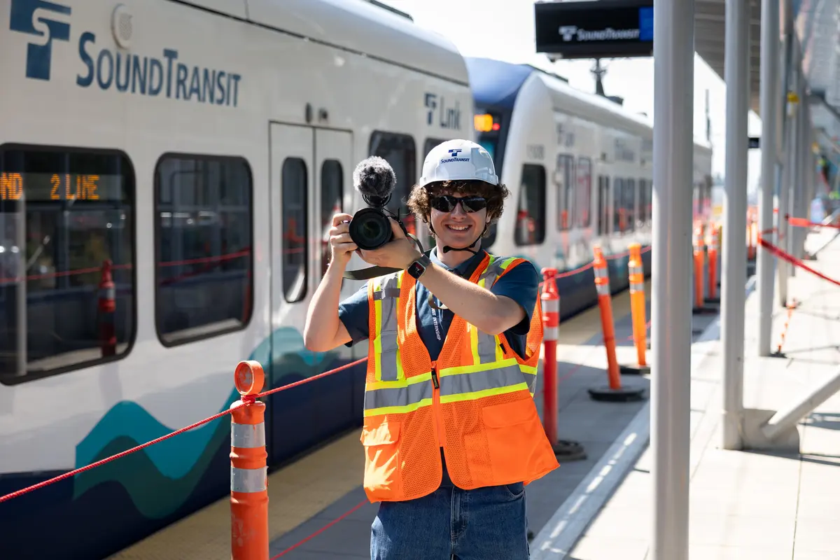 Jonah Kessler-Cohen, wearing a hard hat and safety vest, holds his camera as a light rail passes behind him at the Pinehurst construction site