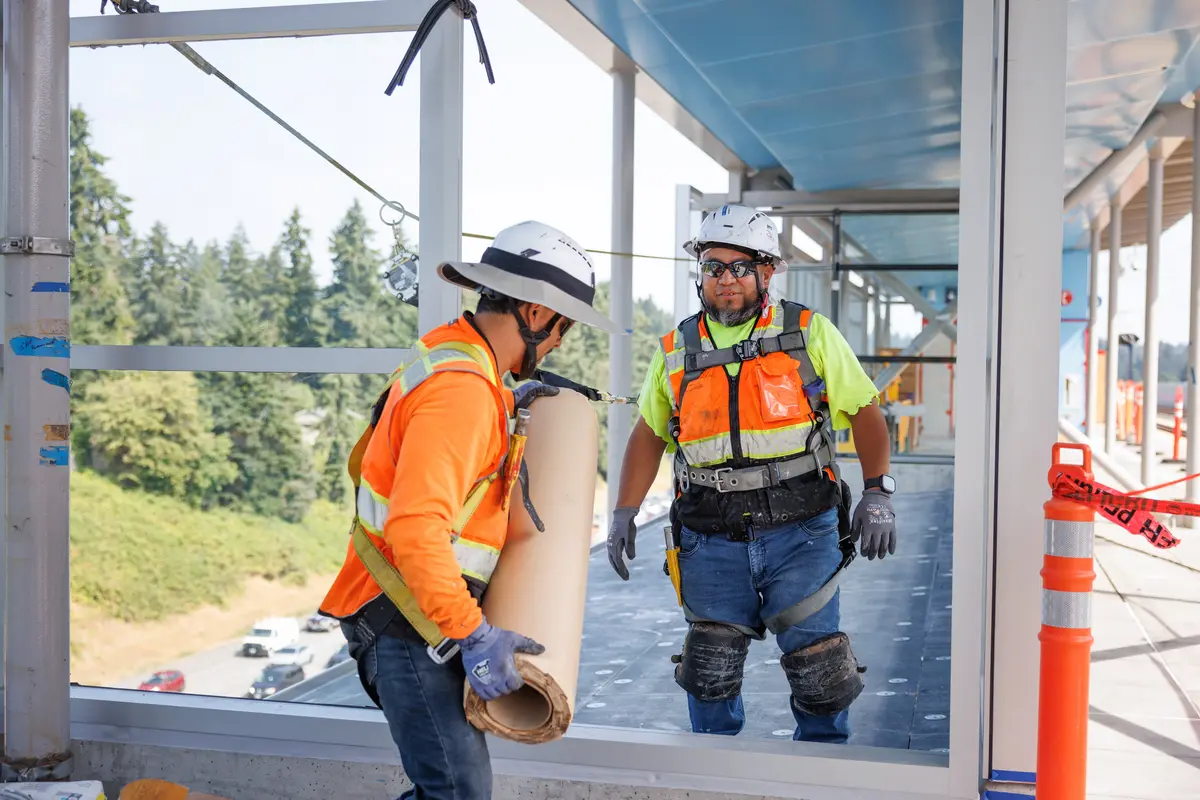 Two harnessed construction workers install roofing at Pinehurst Station